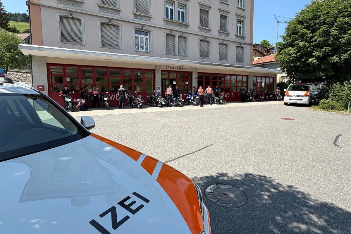 Mehrere Personen stehen vor einem roten Feuerwehrgebäude, daneben Polizeifahrzeuge und Motorräder. Die Szene findet bei Tageslicht statt.