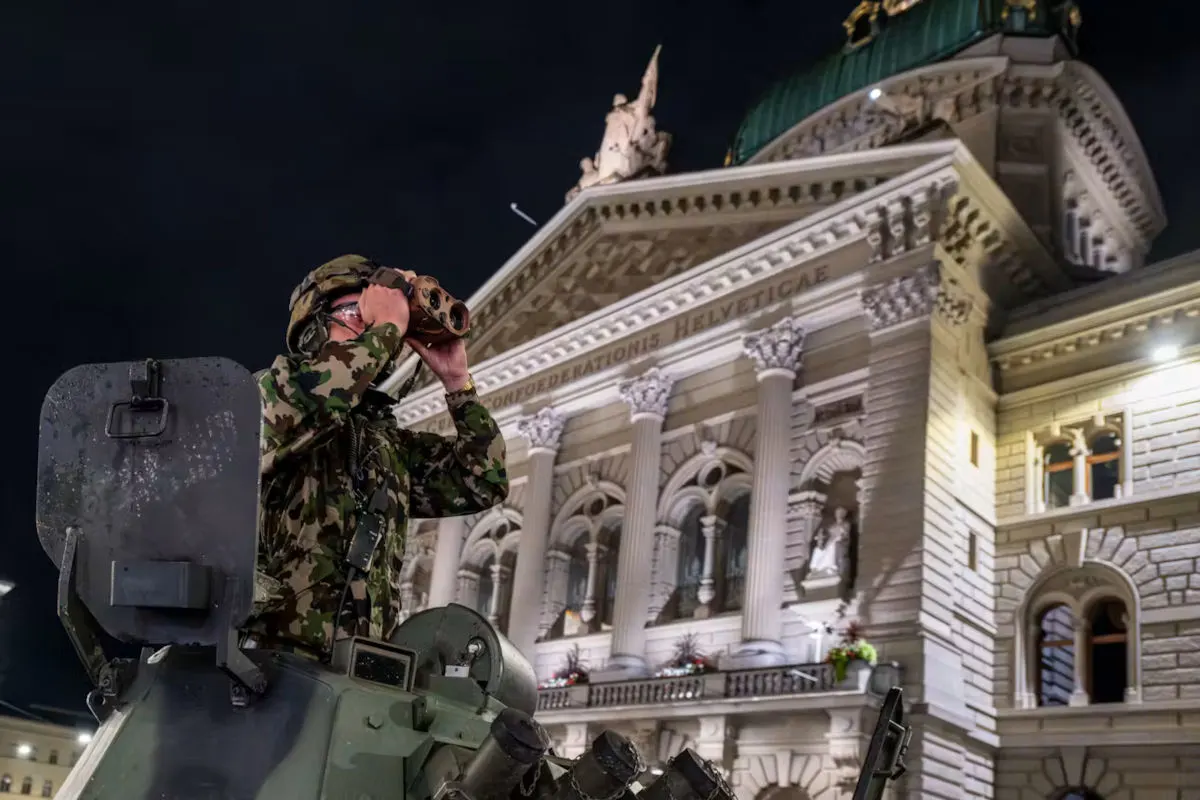 Ein Soldat in Tarnuniform schaut durch ein Fernglas, während er vor dem beleuchteten Bundeshaus in Bern steht.