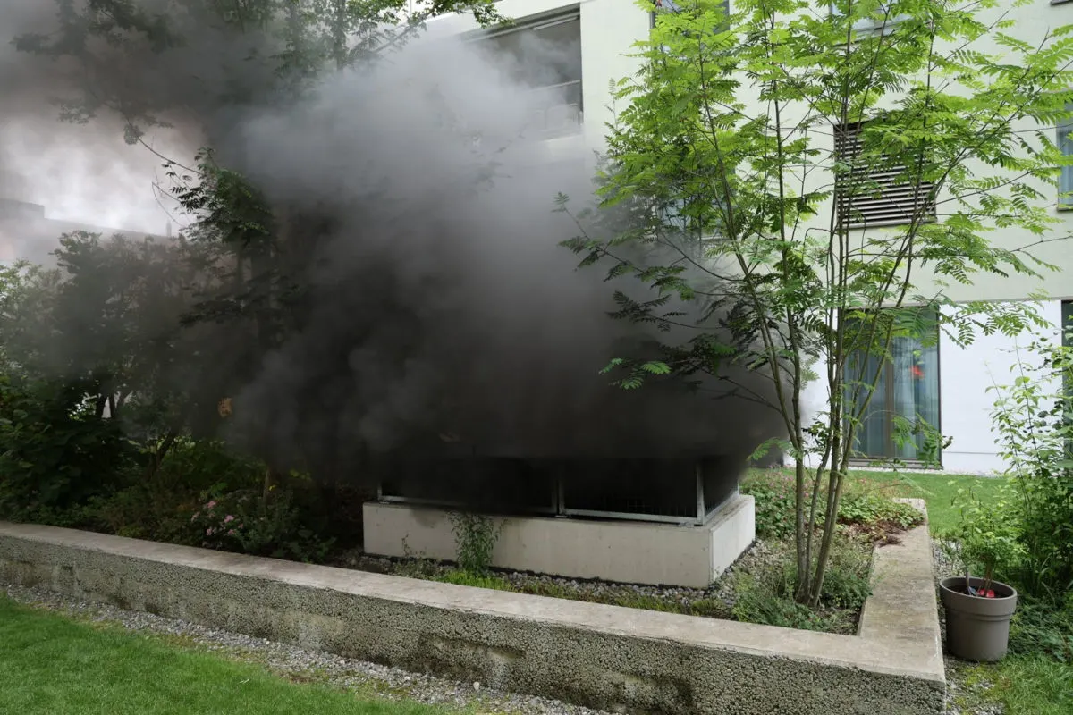 Ein Gebäude in einem Wohngebiet ist von dichtem Rauch umgeben. Der Rauch strömt aus einer Öffnung neben einem Gartenbereich.