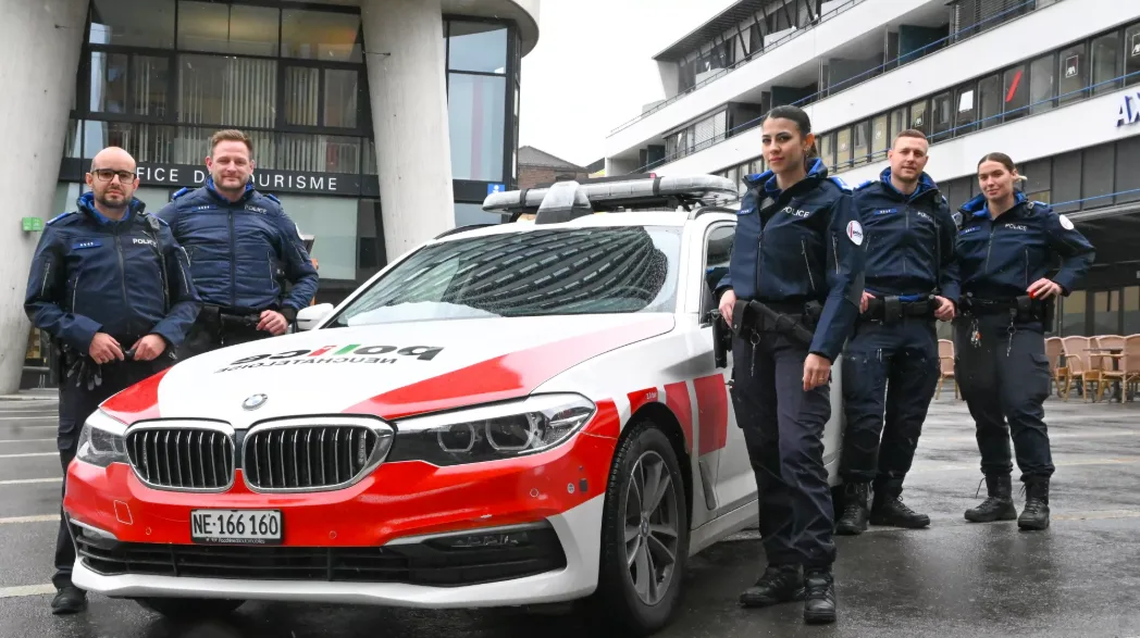 Ein Polizeiwagen der Neuenburger Polizei steht vor einem Gebäude mit der Aufschrift 'Office du Tourisme'. Fünf uniformierte Polizisten stehen um das Fahrzeug.