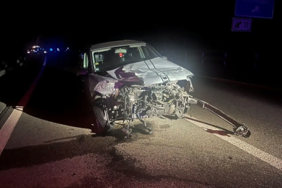 Ein Fahrzeug mit stark zerstörter Frontpartie steht bei Nacht auf einer Autobahn. Im Hintergrund sind Lichter von Einsatzfahrzeugen zu sehen.