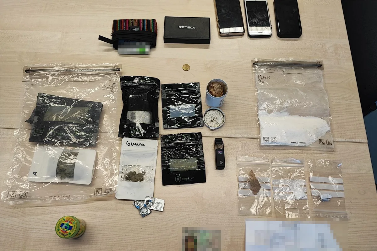 Auf einem Tisch liegen mehrere durchsichtige Tüten mit Substanzen, ein paar Telefone, ein Behälter, eine Münze und weiteres kleines Zubehör. Die Gegenstände wirken wie beschlagnahmt.