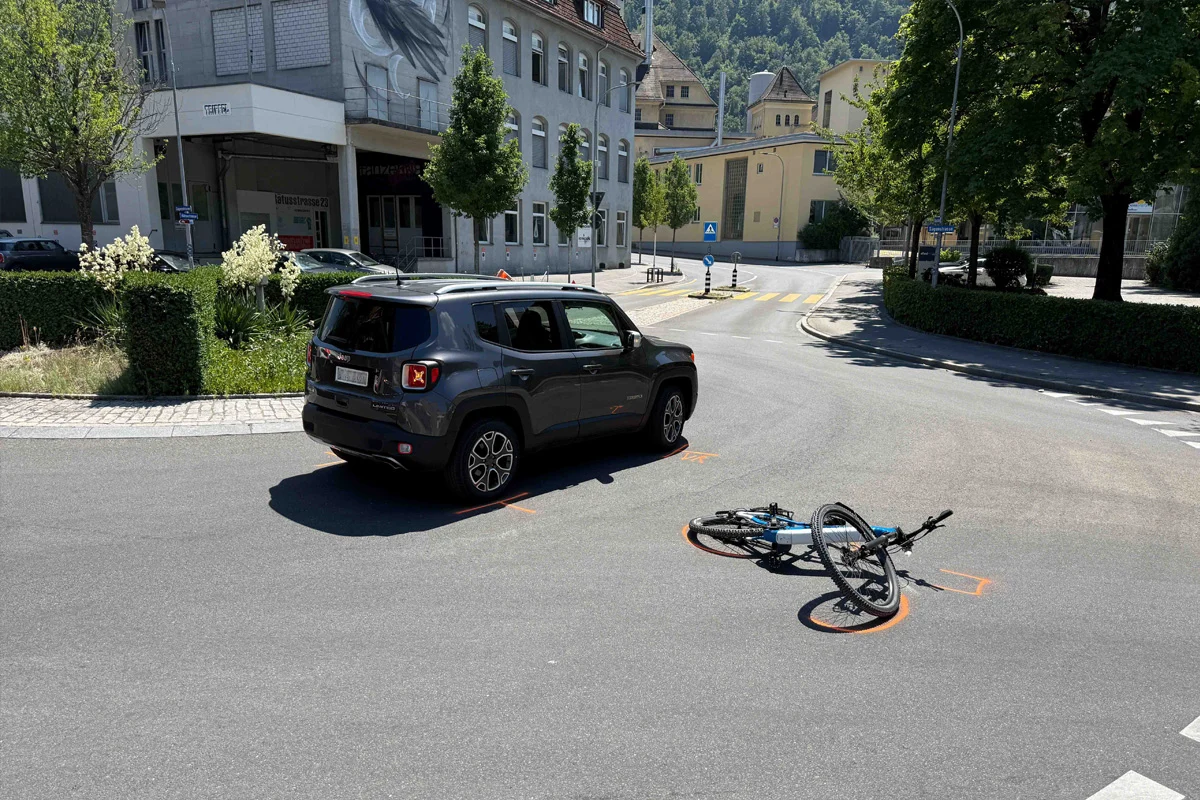 Ein grauer SUV steht auf einer Straße neben einem umgefallenen blauen Fahrrad. Im Hintergrund sind Gebäude und Bäume sichtbar.