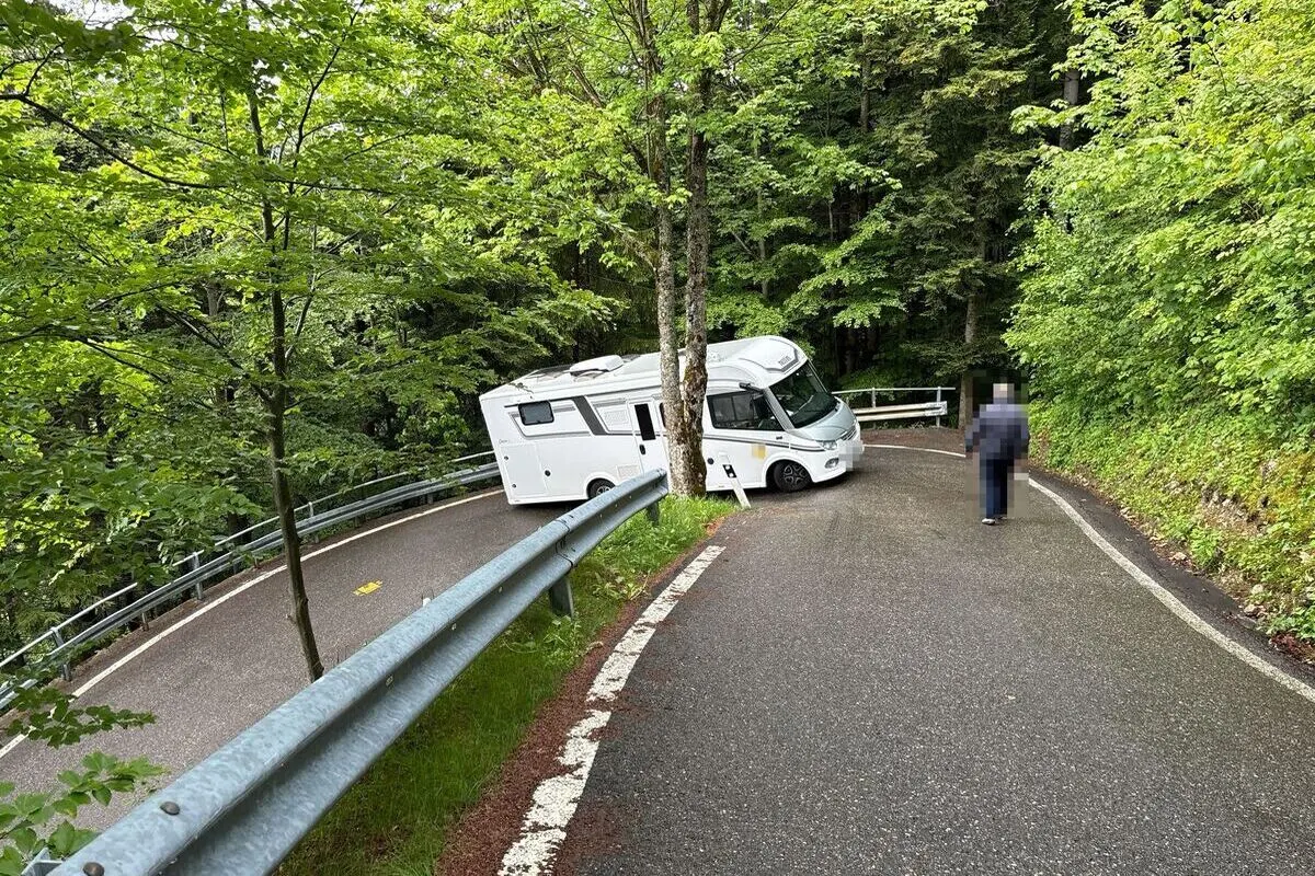 Ein weißes Wohnmobil steckt in einer engen Kurve einer nassen Waldstraße fest. Ein Mensch ist neben dem Fahrzeug sichtbar.
