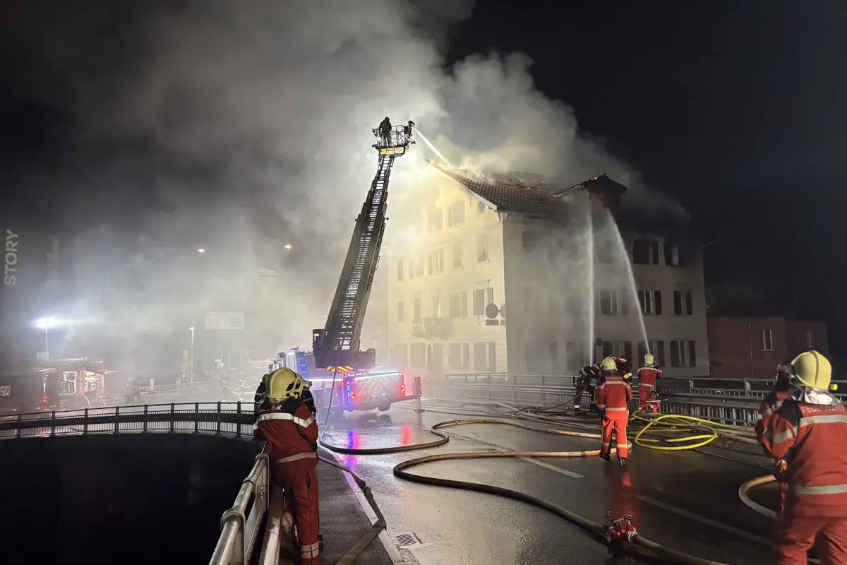 Nachtaufnahme zeigt Feuerwehrleute, die mit Schläuchen einen Dachbrand bekämpfen. Ein Leiterfahrzeug unterstützt die Löschaktion.
