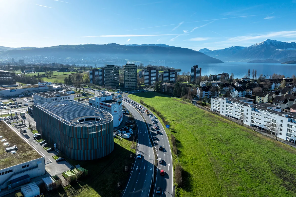 Luftaufnahme zeigt eine Stadt mit einigen Hochhäusern in der Nähe eines Sees, umgeben von grünen Flächen und Bergen im Hintergrund. Mehrere Autos sind auf einer engen Kurve der Straße zu sehen.