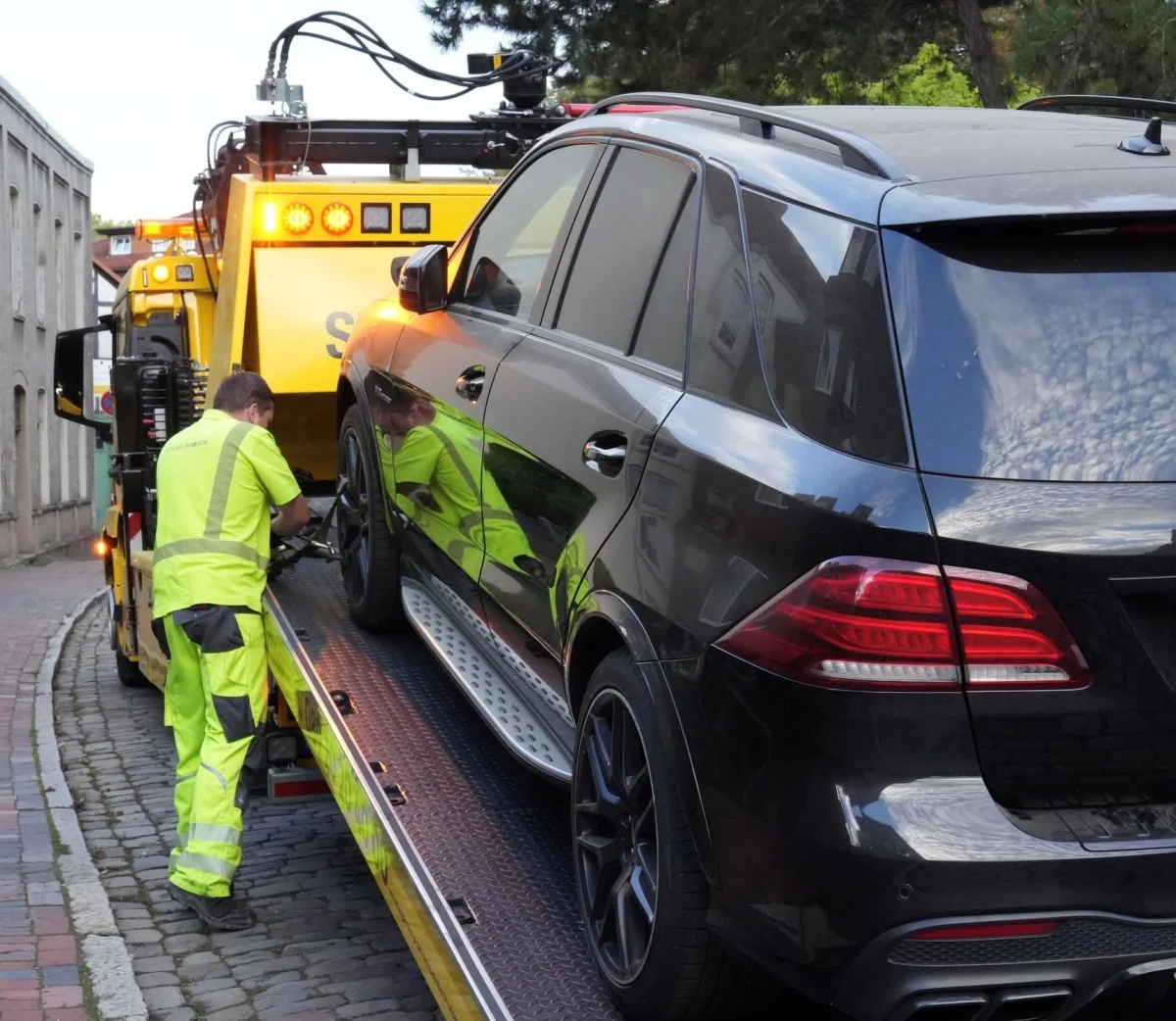 Ein Abschleppwagen lädt einen dunklen SUV auf einer gepflasterten Straße. Eine Person in Arbeitskleidung sichert das Fahrzeug.