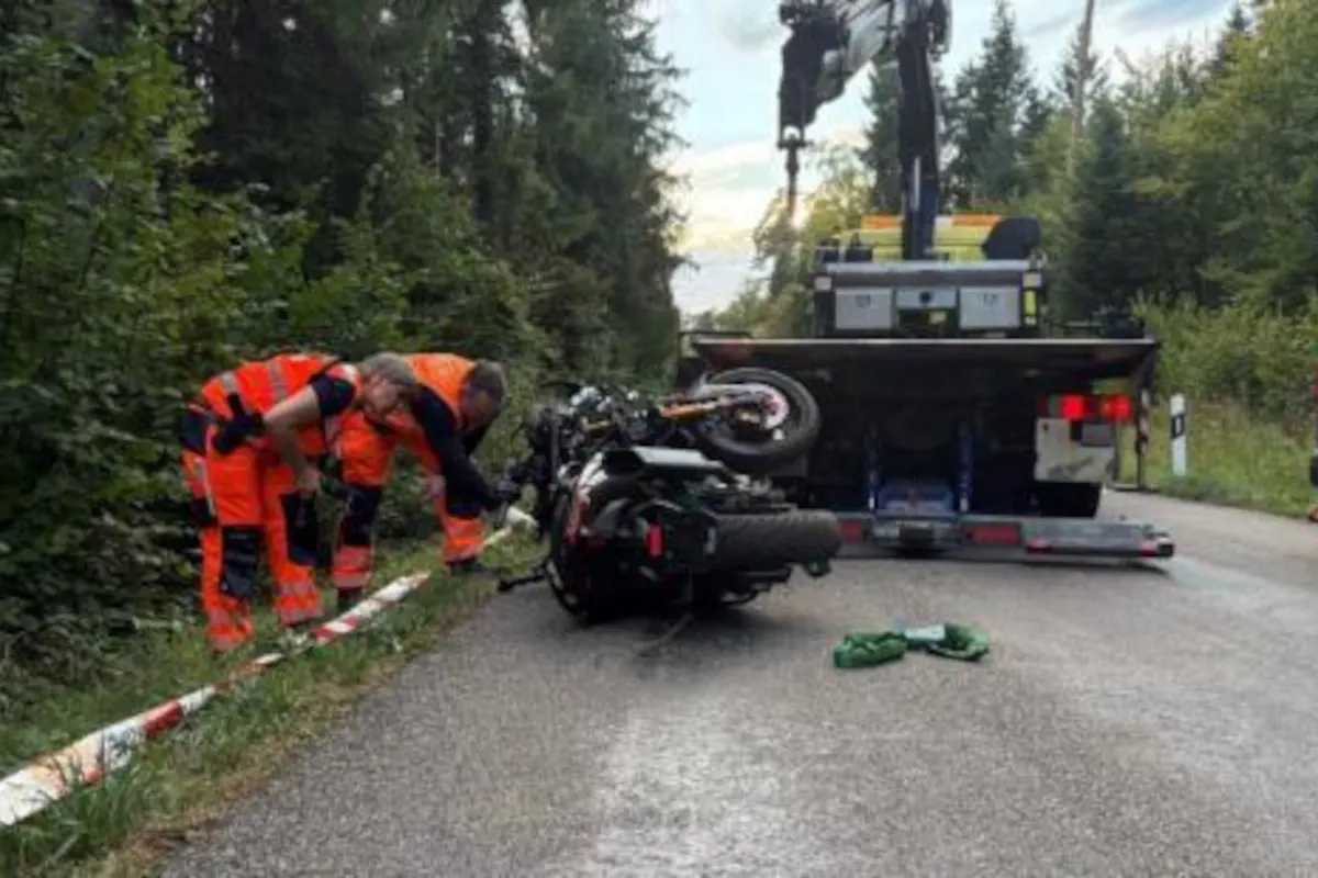 Zwei Personen tragen orangefarbene Warnwesten und untersuchen ein umgestürztes Motorrad auf einer bewaldeten Straße, im Hintergrund ist ein Abschleppfahrzeug zu sehen.