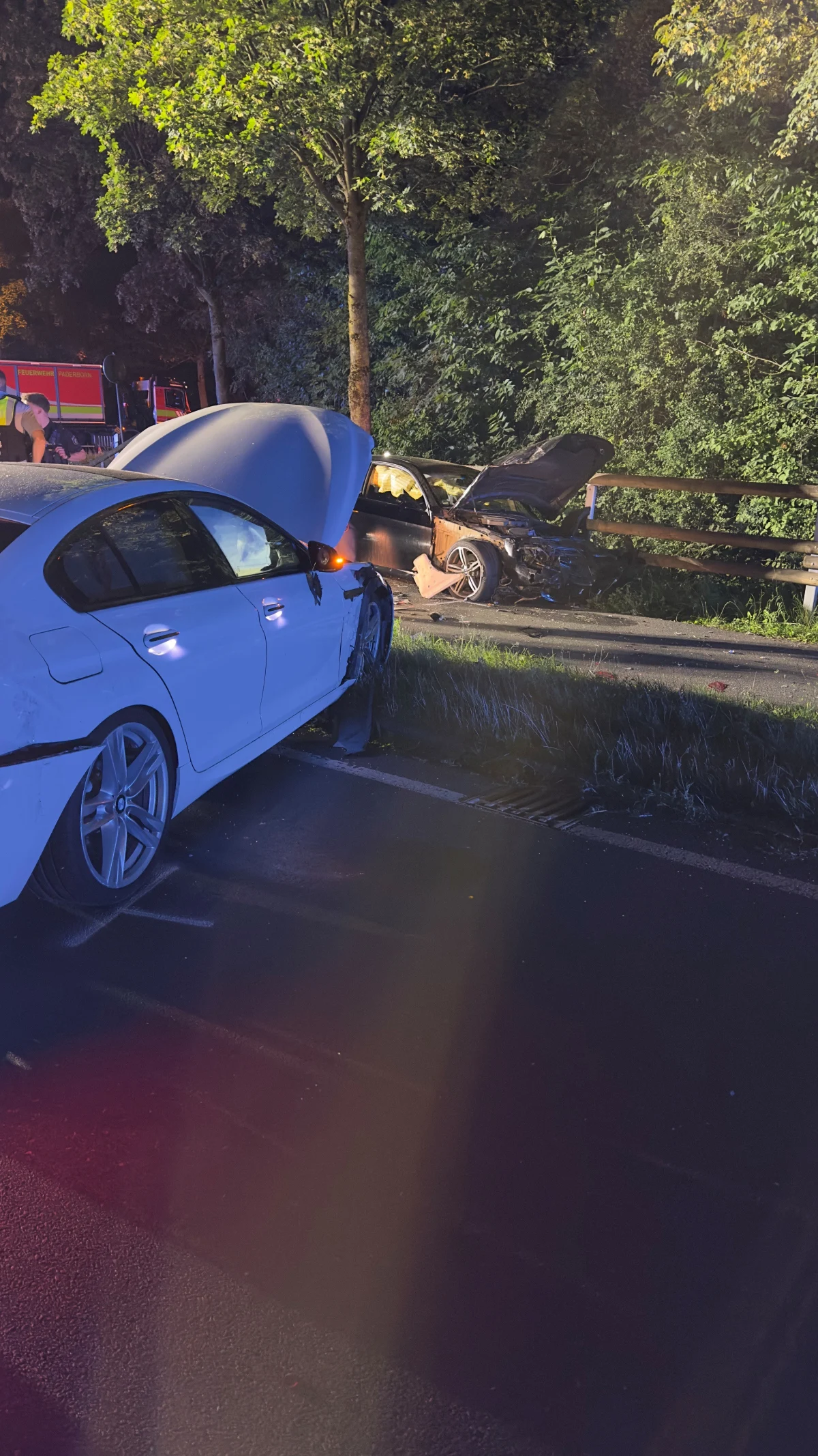Im Vordergrund ein weißes Auto mit starkem Frontschaden, im Hintergrund ein weiteres Auto mit geöffnetem Kofferraum, ebenfalls beschädigt.