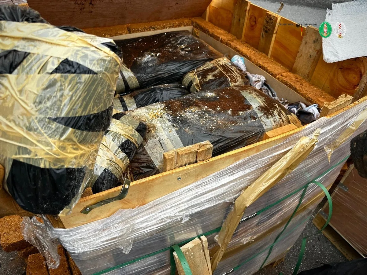 Eine geöffnete Holzkiste zeigt mehrere schwarze, in Plastik verpackte Pakete. Die Verpackung ist teilweise mit Klebeband umwickelt. Umgebung deutet auf eine industrielle oder logistische Szene hin.
