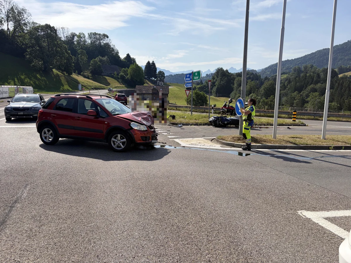Ein roter PKW steht mit beschädigter Front auf einer Straße, daneben ein Arbeiter in Warnkleidung mit einem Verkehrsschild.