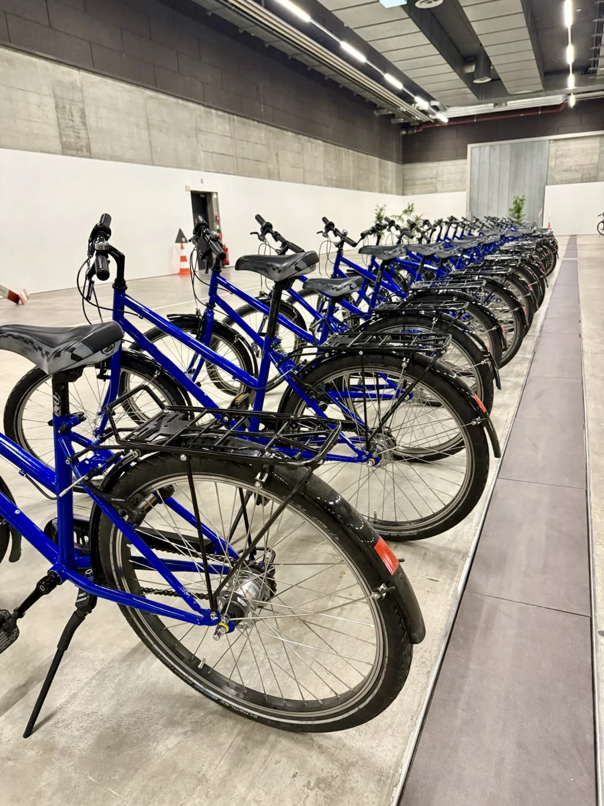 In einem modernen Innenraum steht eine lange Reihe blauer Fahrräder ordentlich nebeneinander. Der Hintergrund ist eine Betonwand.