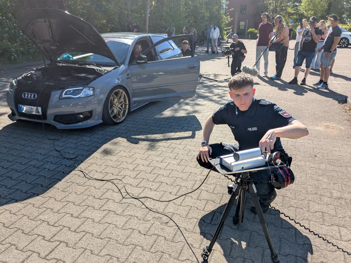 Ein Polizist ist vor einem grauen Audi mit geöffneter Motorhaube und führt Messungen durch. Mehrere Menschen stehen im Hintergrund.