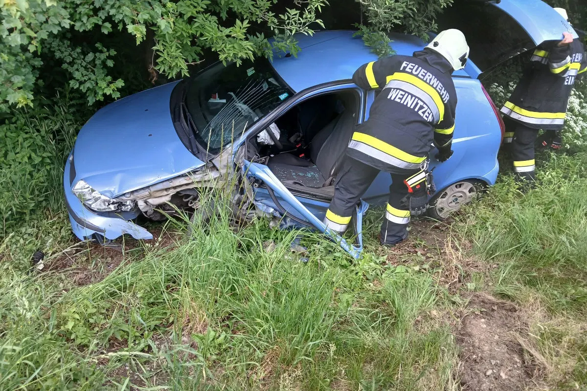 Ein blaues Auto liegt stark beschädigt im Graben eines bewachsenen Bereichs. Zwei Feuerwehrleute von Weinitzen arbeiten am Fahrzeug, um es zu bergen.