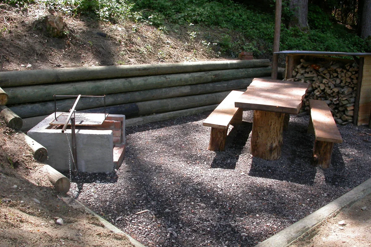 Ein Grillplatz mit einer Holzbank, einem Tisch und einem Stapel Feuerholz neben der Grillstelle im Wald.
