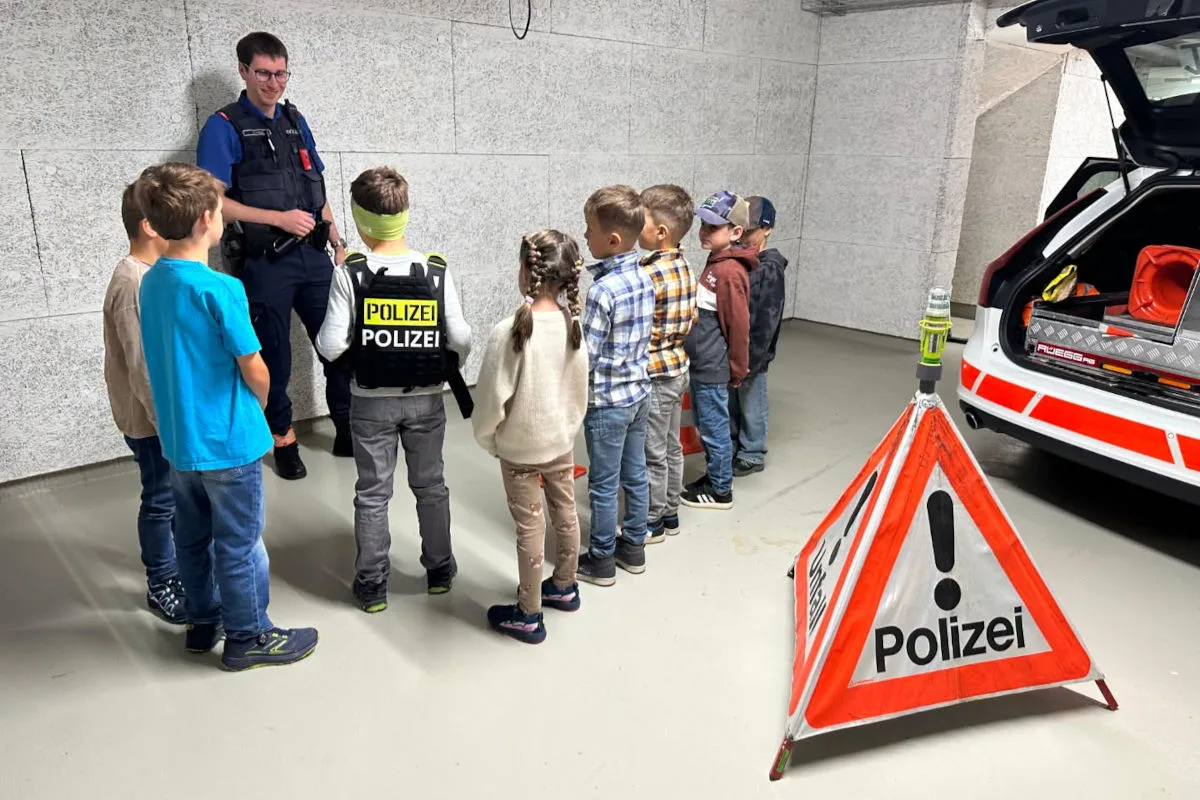 Ein Polizist hält eine Schulung für eine Gruppe von Kindern in einer Garage. Ein Polizeiwagen mit geöffnetem Kofferraum steht daneben.