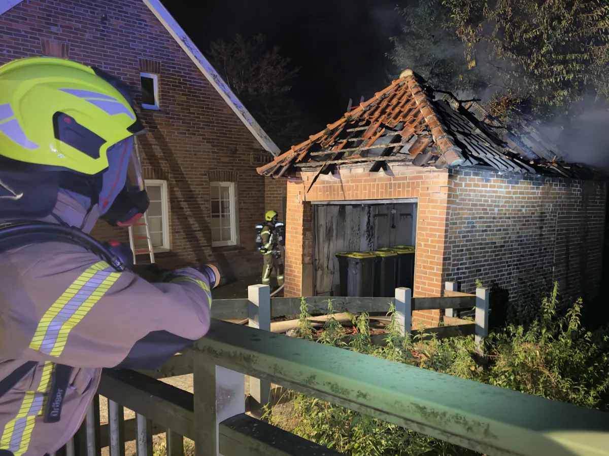 Zwei Feuerwehrleute in voller Ausrüstung bekämpfen nachts einen Brand an einem Backsteinschuppen. Rauch steigt vom Dach auf.