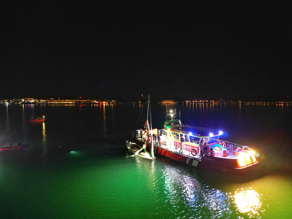 Ein rot-weißes Feuerwehrboot mit eingeschaltetem Blaulicht ist bei Nacht auf einem See im Einsatz. Menschen sind auf dem Deck des Bootes sichtbar, und ein gesunkenes Boot wird geborgen.
