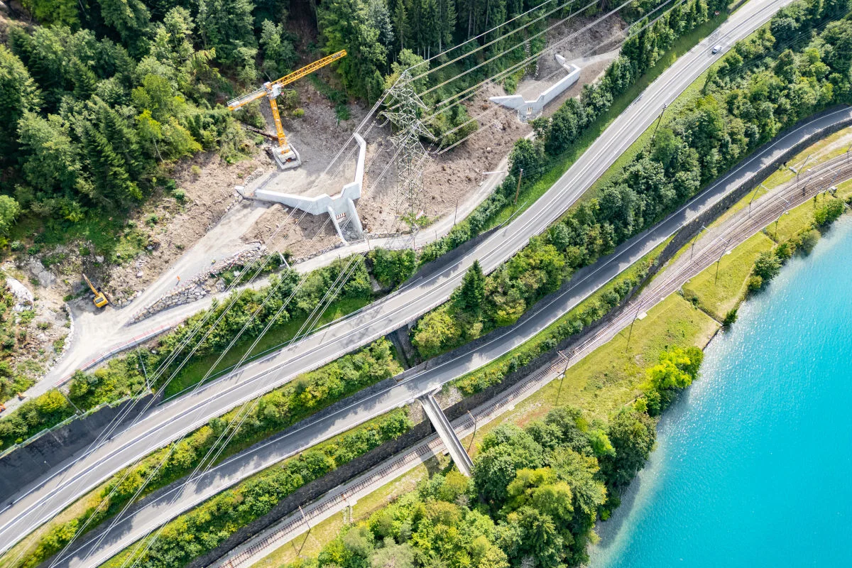 Eine Luftaufnahme zeigt eine Baustelle mit einem Kran in einem Waldgebiet neben einer Straße und Stromleitungen. Der Bereich wird durch Straßen und Schienen begrenzt, mit einem Fluss rechts im Bild.