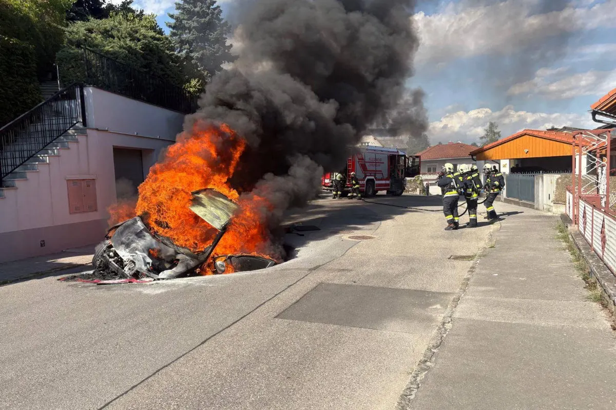 Ein Auto steht auf der Straße in Flammen, während Feuerwehrleute in Schutzkleidung versuchen, den Brand zu löschen. Schwarzer Rauch steigt in den Himmel.