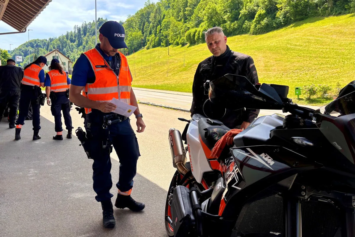 Ein Polizist in Warnweste kontrolliert die Dokumente eines Motorradfahrers. Im Hintergrund sind weitere Polizisten zu sehen.