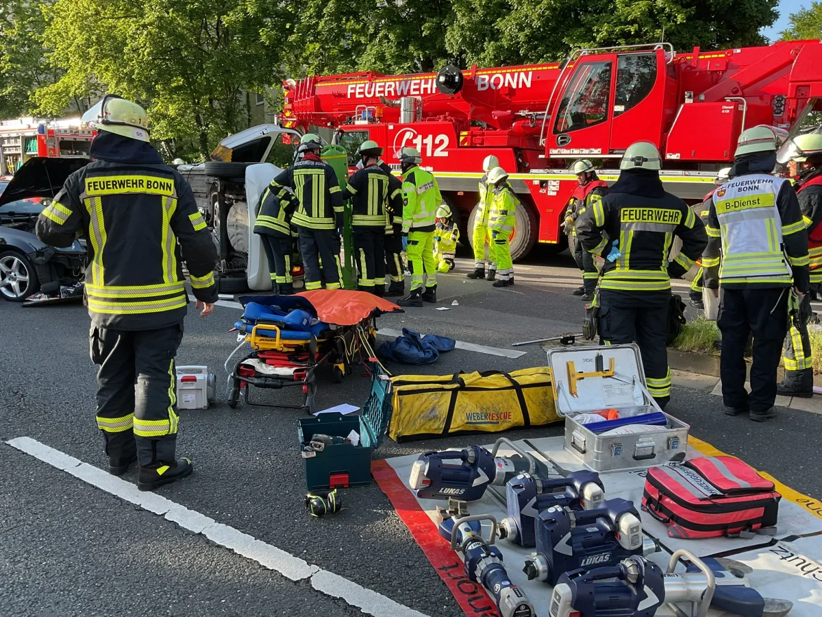 Mehrere Feuerwehrleute, gekennzeichnet als Feuerwehr Bonn, arbeiten mit Rettungsgeräten an einem umgestürzten Auto auf einer Straße. Im Hintergrund ist ein Feuerwehrfahrzeug zu sehen.
