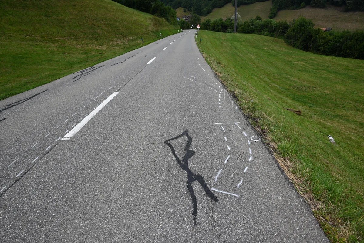 Die Straße zeigt schwarze und weiße Markierungen, die auf einen Unfall hinweisen. Sie verläuft durch eine grüne Landschaft.