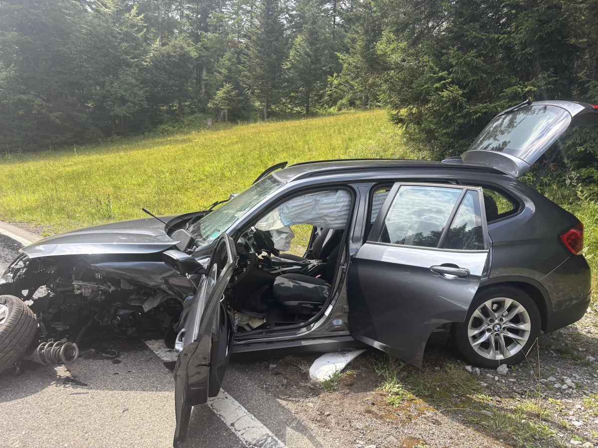 Ein stark beschädigter, dunkler BMW steht am Straßenrand mit offenem Kofferraum. Die Umgebung zeigt eine Waldlandschaft.
