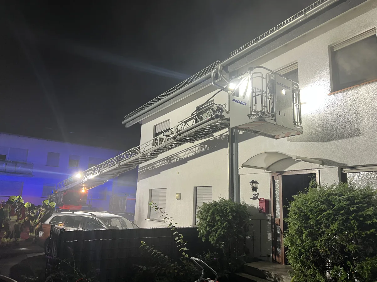 Eine Feuerwehrleiter ist an einem zweistöckigen Gebäude angelehnt, und mehrere Feuerwehrleute stehen in der Nähe eines Einsatzfahrzeugs bei Nacht.