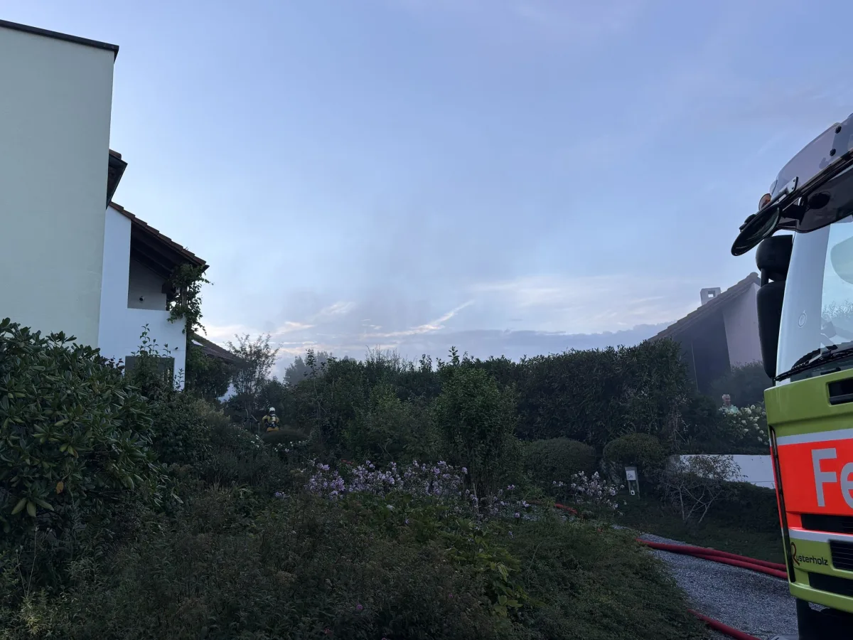 Ein Feuerwehrfahrzeug steht in der Nähe eines Wohnhauses, während ein Feuerwehrmann im Garten sichtbar ist. Rauch ist im Hintergrund zu sehen.