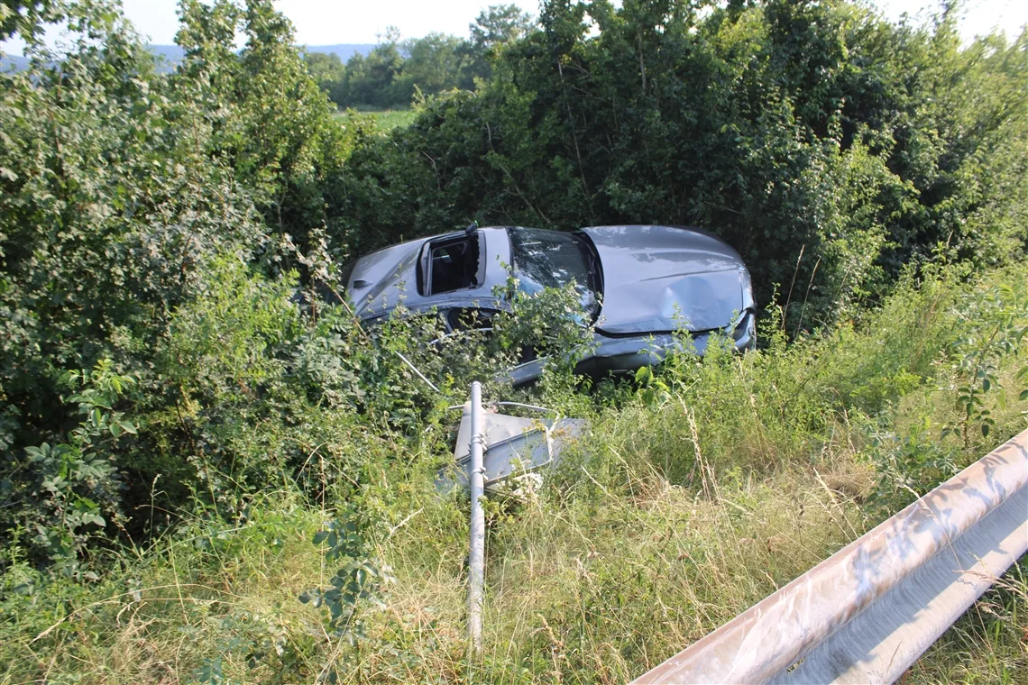 Ein silbernes Auto ist von der Straße abgekommen und liegt im dichten Gebüsch. Vor dem Auto ist ein verbogenes Verkehrsschild zu sehen.