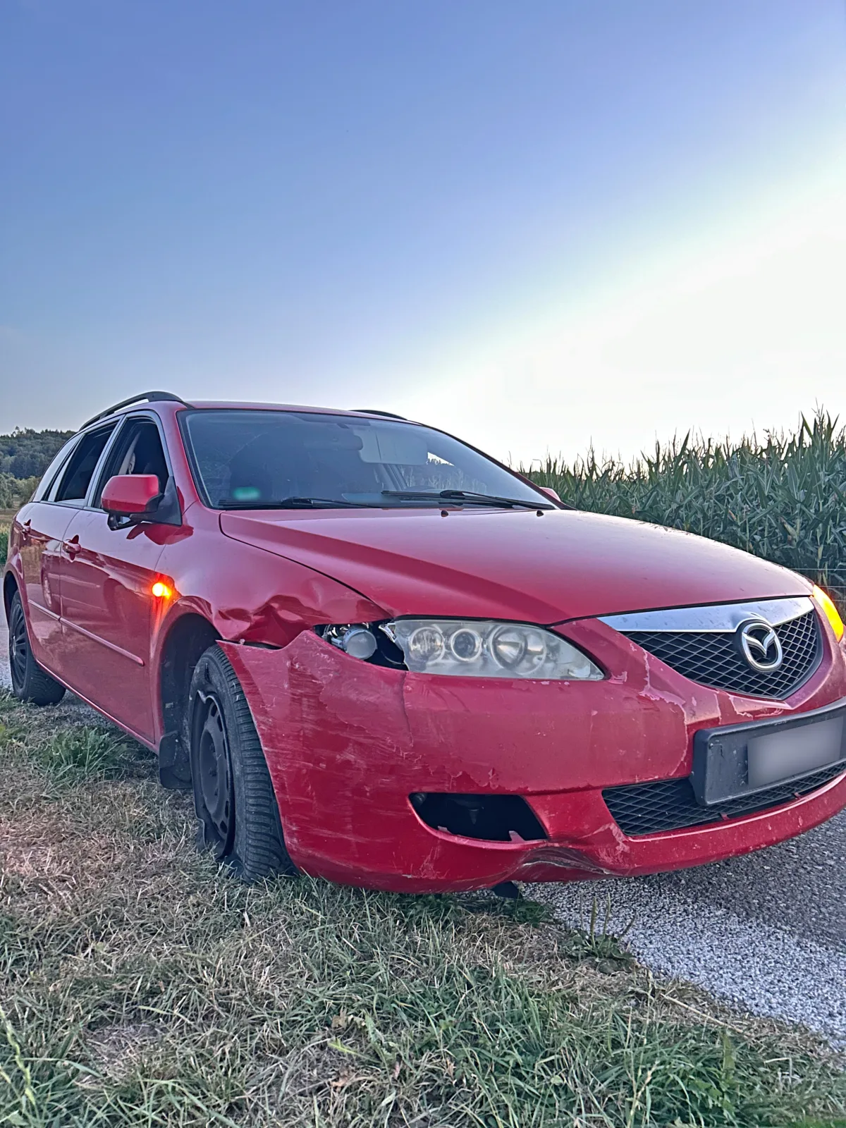 Ein roter Mazda-Kombi mit Frontschäden steht am Straßenrand neben einem Feld. Es ist ein klarer Tag.