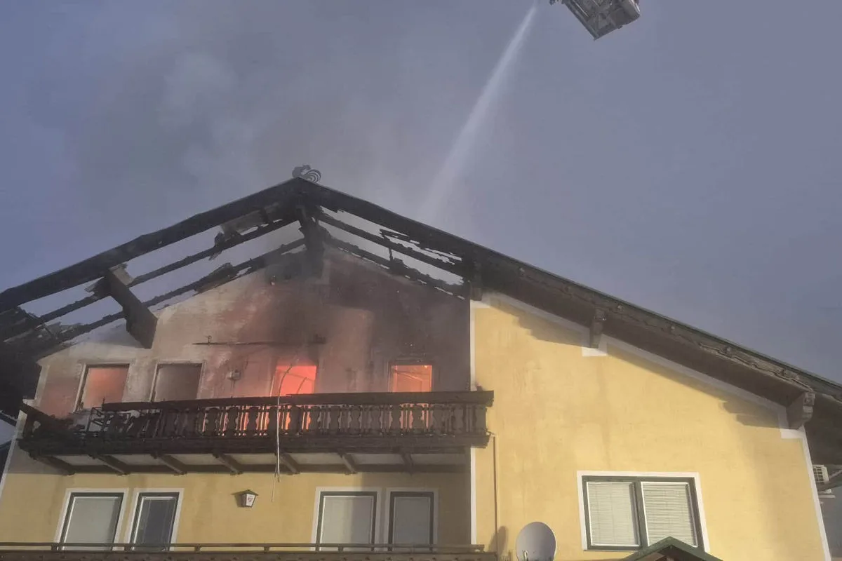 Ein gelbes Haus zeigt einen brennenden Dachstuhl, während die Feuerwehr das Feuer löscht. Flammen sind sichtbar.