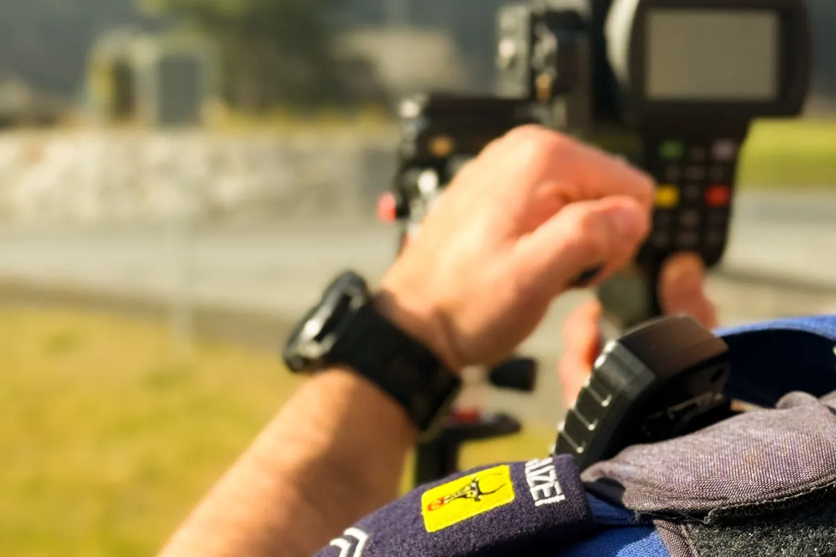 Ein Polizeibeamter mit Armbanduhr hält ein Geschwindigkeitsmessgerät. Im Vordergrund ist ein Polizeiemblem sichtbar.