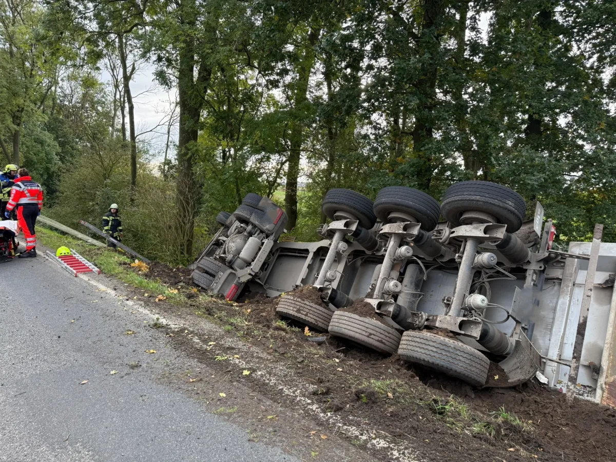 Ein Lastwagen liegt umgekippt im Graben neben einer Landstraße, während Feuerwehrleute an der Unfallstelle tätig sind.