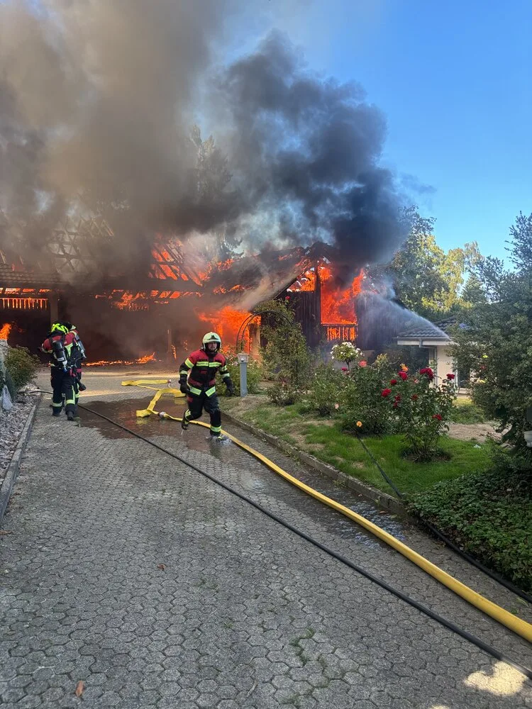 Mehrere Feuerwehrleute in Schutzkleidung bekämpfen einen großen Brand in einem Holzgebäude, während dunkler Rauch aufsteigt.