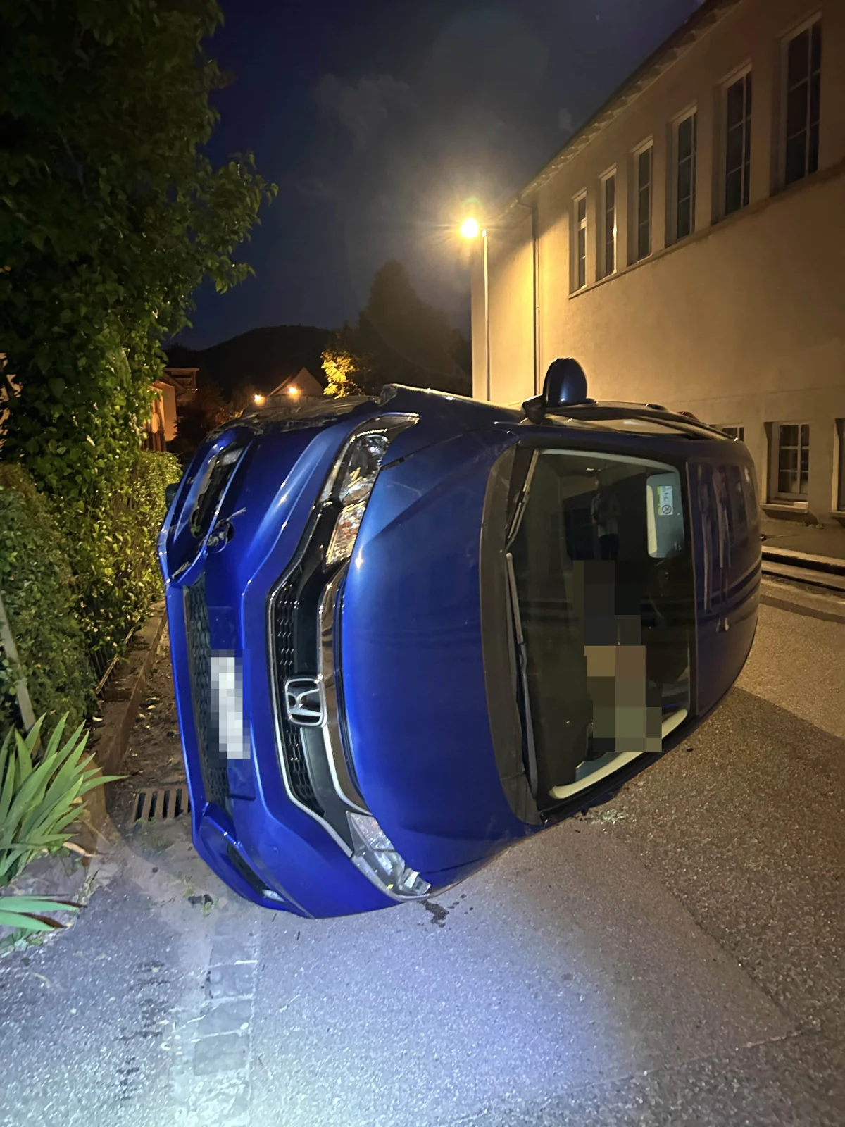 Ein blaues Auto ist auf die Seite gekippt neben einer Straße. Es ist Nacht, und Straßenlaternen beleuchten die Szene.