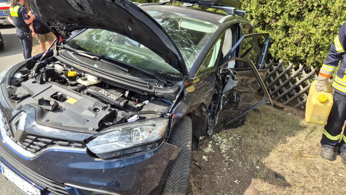 Ein stark beschädigtes Auto mit geöffneter Motorhaube steht neben einem umgestürzten Holzzaun. Eine Person in Uniform hält einen Kanister.