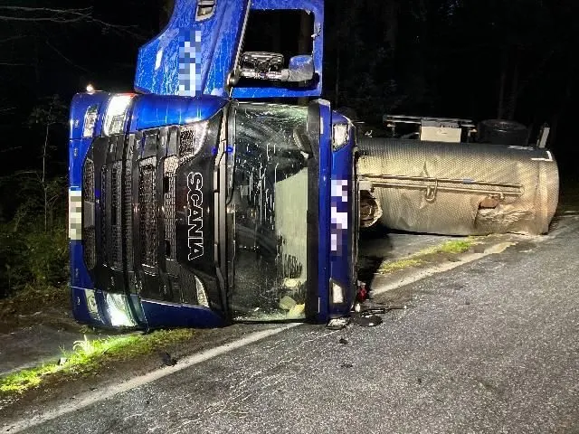 Ein großer blauer Lastwagen liegt seitlich auf einer abgelegenen, dunklen Straße. Der Asphalt ist durch den Unfall leicht beschädigt.