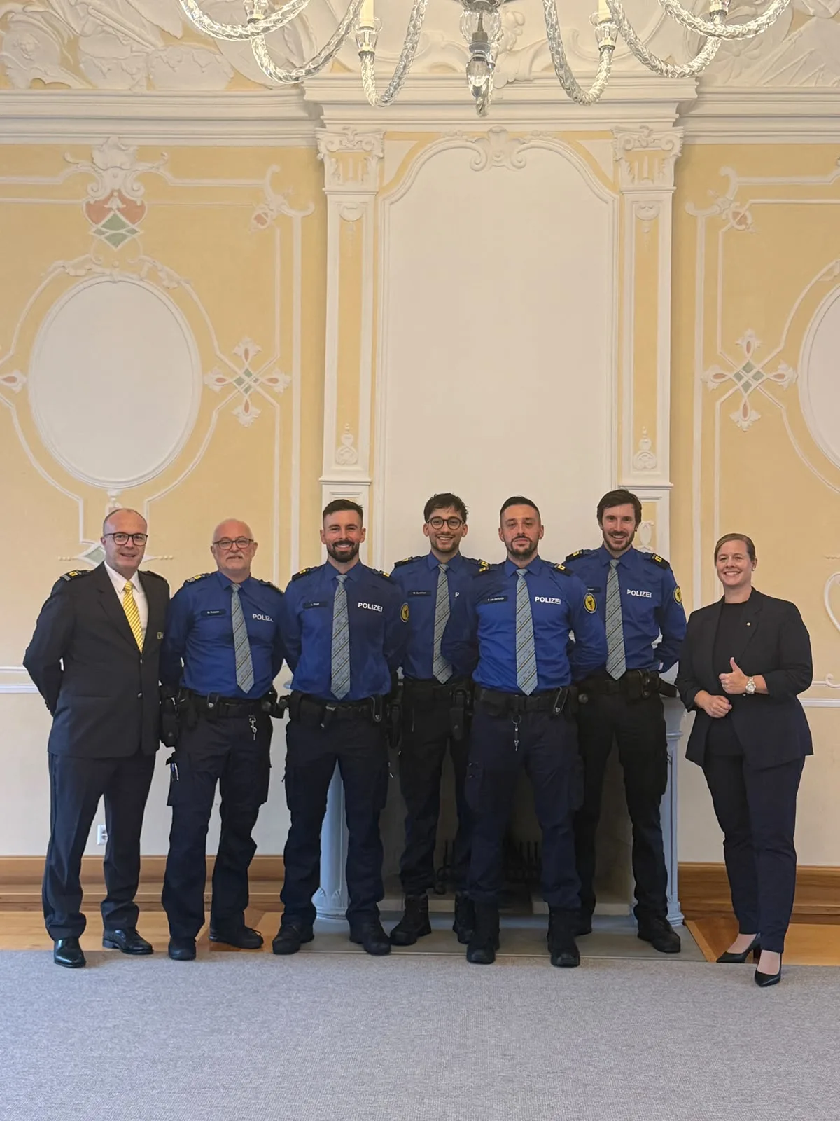 Eine Gruppe von sieben Personen, darunter Polizisten in blauer Uniform und eine Frau in Zivil, steht vor einer dekorierten Wand in einem Innenraum.