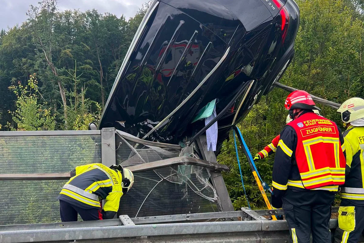 Ein schwarzes Auto ist auf einer Leitplanke aufgebockt, während mehrere Feuerwehrleute in Ausrüstung daran arbeiten. Im Hintergrund ist Wald zu sehen.