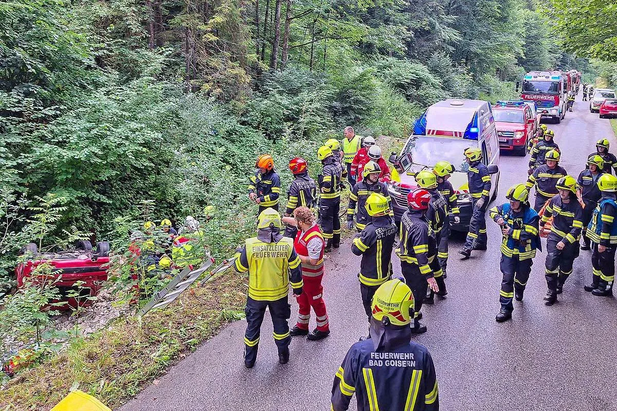 Feuerwehrkräfte stehen in einem Waldgebiet an einem umgestürzten Auto am Hang. Mehrere Einsatzfahrzeuge sind auf der Straße.