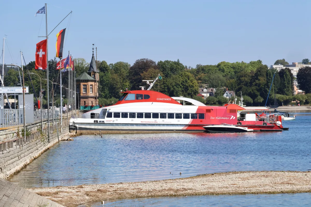 Ein Katamaran mit der Aufschrift 'Constanze' liegt am Ufer eines Sees. Mehrere nationale Fahnen sind gehisst.