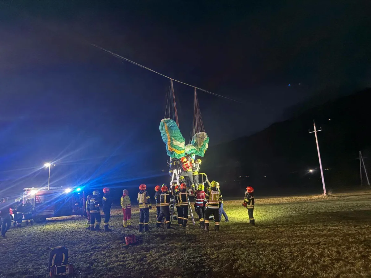 Bei Nacht befreien mehrere Rettungskräfte einen Paraglider, der in Stromleitungen auf einem Feld hängt. Ein Einsatzfahrzeug ist am Rand des Feldes zu sehen.