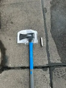 Ein verpackter Hammer mit blauem Griff und eine kleine Patronenhülse liegen auf einem Gehweg aus Pflastersteinen.