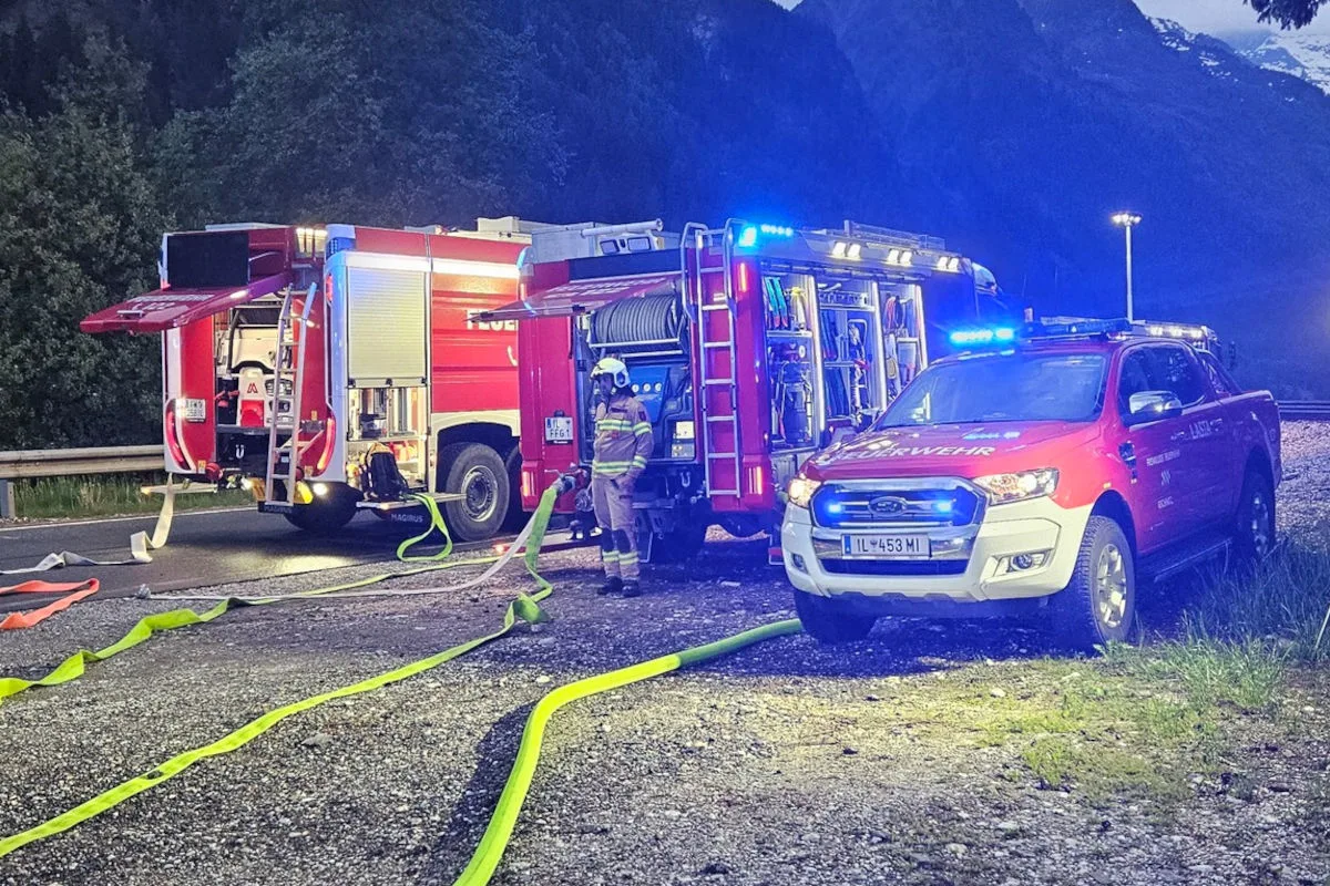 Zwei rote Feuerwehrfahrzeuge stehen mit ausgerollten Schläuchen auf einer Straßenfläche, umgeben von Bergen im Hintergrund. Ein Feuerwehrmann in Uniform ist sichtbar.