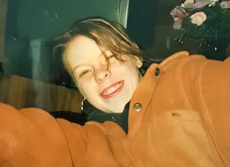 Eine lächelnde Person trägt einen orangefarbenen Mantel. Im Hintergrund sind Pflanzen sichtbar, was auf einen Innenraum hinweist.