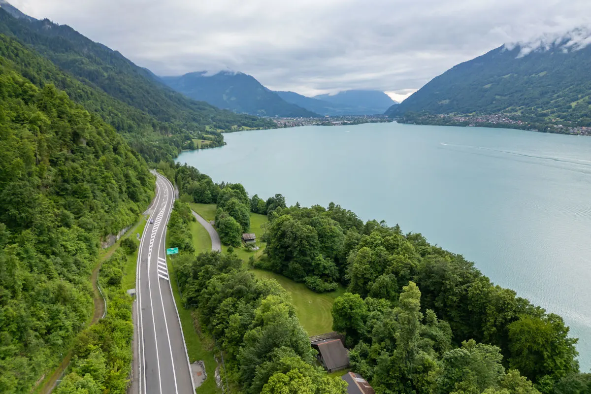 Eine Straße schlängelt sich am Ufer eines großen Sees entlang, umgeben von grünen Hügeln und Bergen.