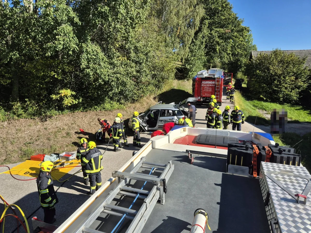 Mehrere Feuerwehrleute in Schutzkleidung arbeiten an einem verunfallten Auto auf einer Landstraße. Im Hintergrund sind Feuerwehrfahrzeuge sichtbar.
