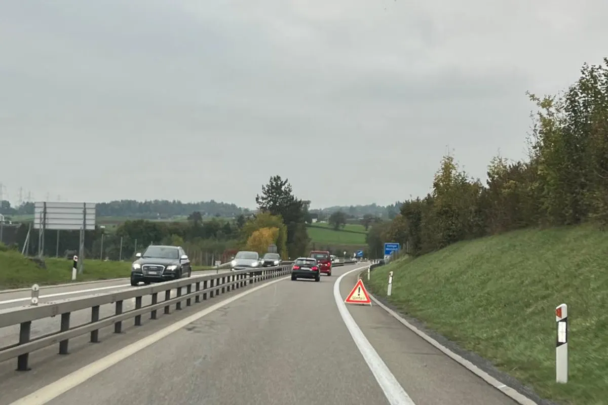 Mehrere Fahrzeuge fahren auf einer Autobahn, die durch ein Verkehrszeichen 'Unfall' am rechten Rand gekennzeichnet ist. Der Himmel ist bewölkt und die Umgebung ist grün mit Bäumen.